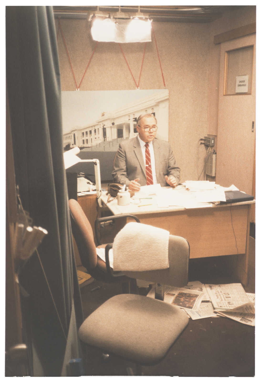 Laurie Oakes sitting at a desk in front of a held up image of Old Parliament House on Camera with pieces of paper scattered all over his desk.