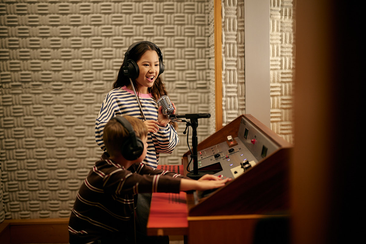 Two kids in a makeshift radio booth, interacting with a sound board and microphone wearing headphones.