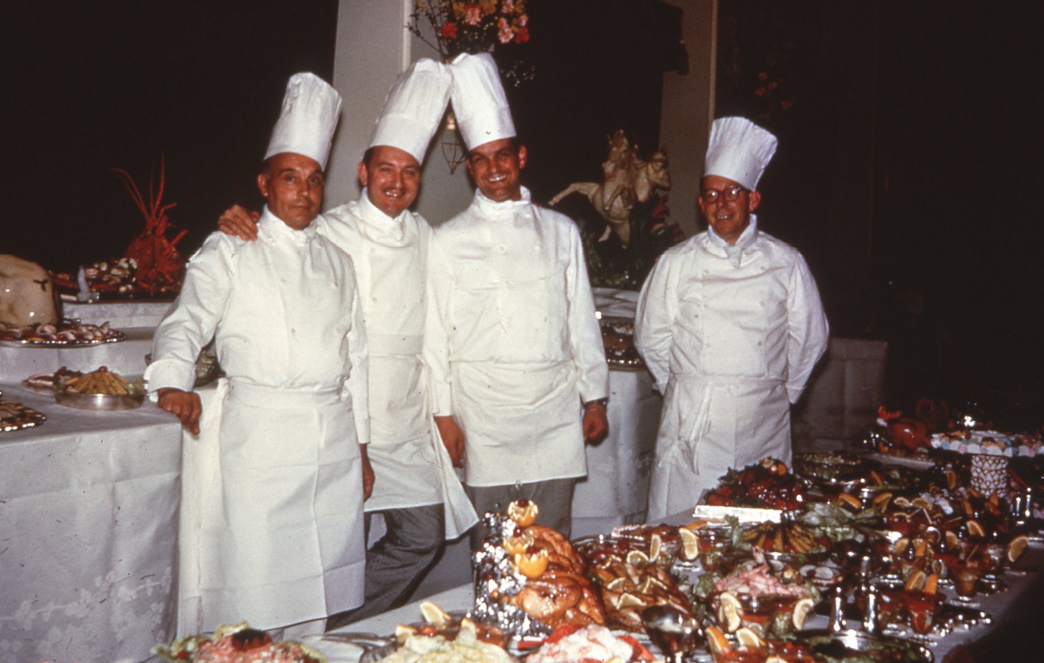 Four chefs standing in the dining room surrounded by banquet tables covered in food.