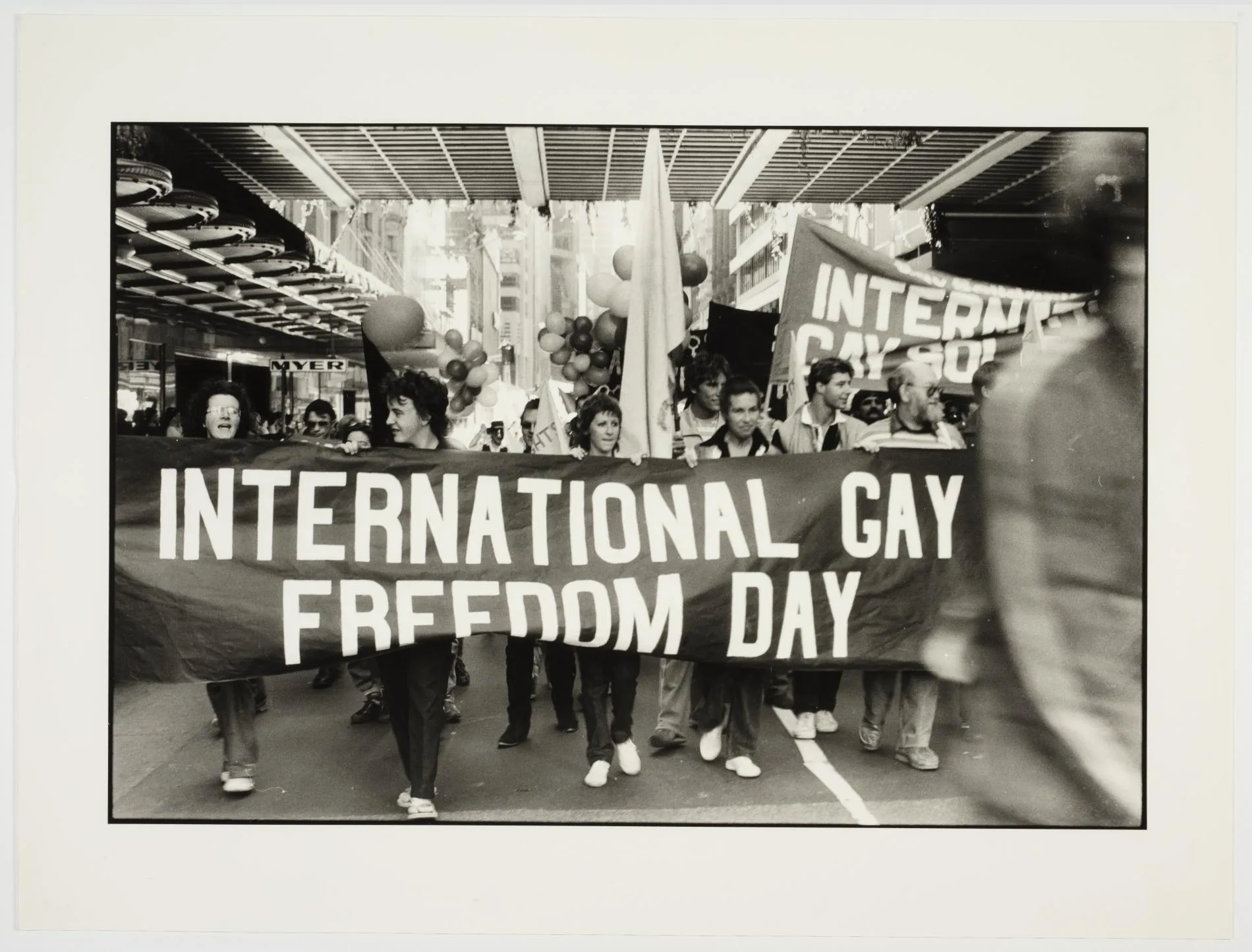 A black and white image of a protest during Stonewall Week in 1981. Prominently pictured are some people holding up a large sheet sign that says 'International Gay Freedom Day'.