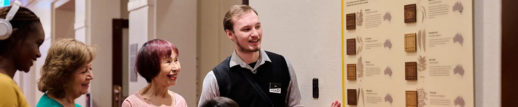 A Museum staff member and a group of people in brightly coloured tops stand in a hallway with red carpet looking at a board displaying Australian timbers used in Old Parliament House.