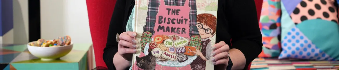 A woman sitting holding a picture book titled 'The Biscuit Maker' by Sue Lawson, illustrated by Liz Anelli