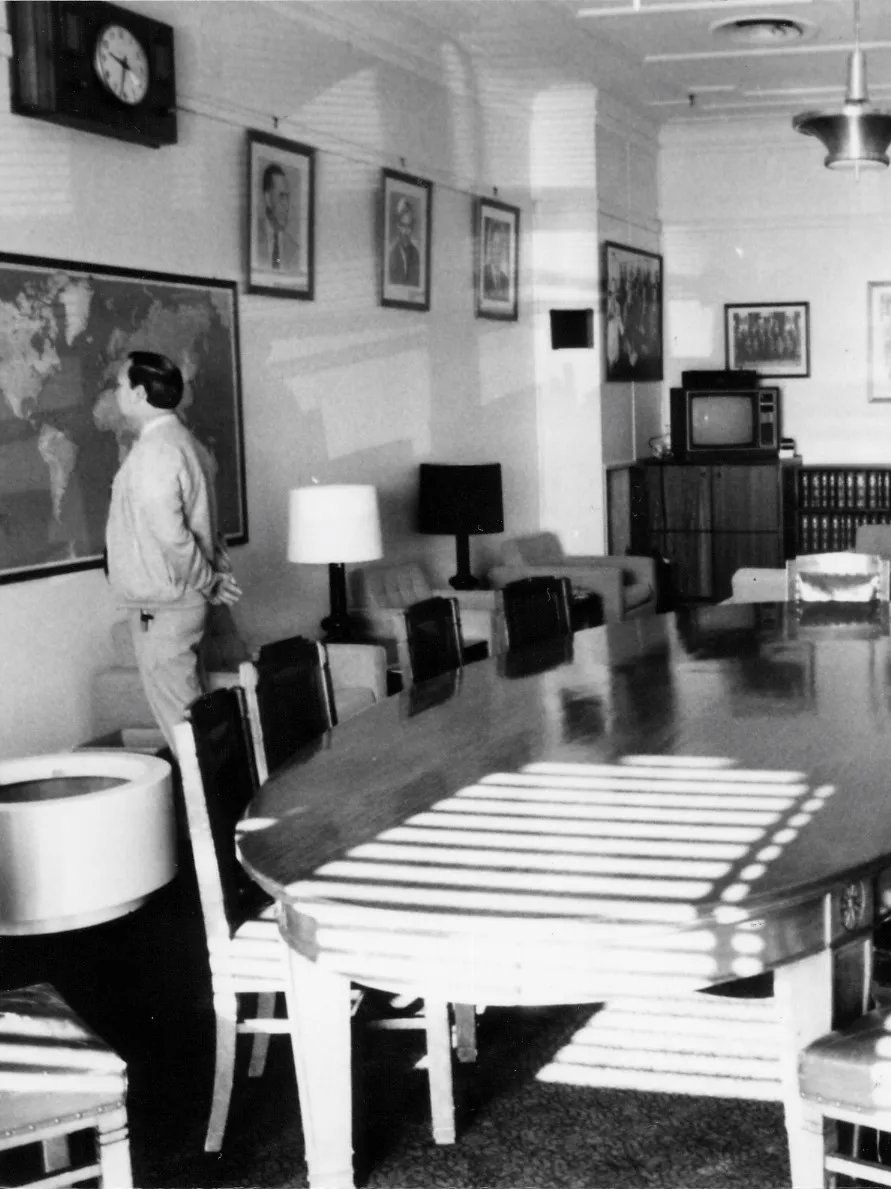 A large oval meeting table and chairs dominate the centre of the room. Easy chairs with side tables and lamps are arrayed along the sides and two sofas are arranged in a seating area towards the back of the room. A sideboard with books, a television and sound system stretches along the back wall. There is a framed map on one wall and a notice board on the opposite. Portraits of leaders and formal and informal photographs of the party crowd the walls
