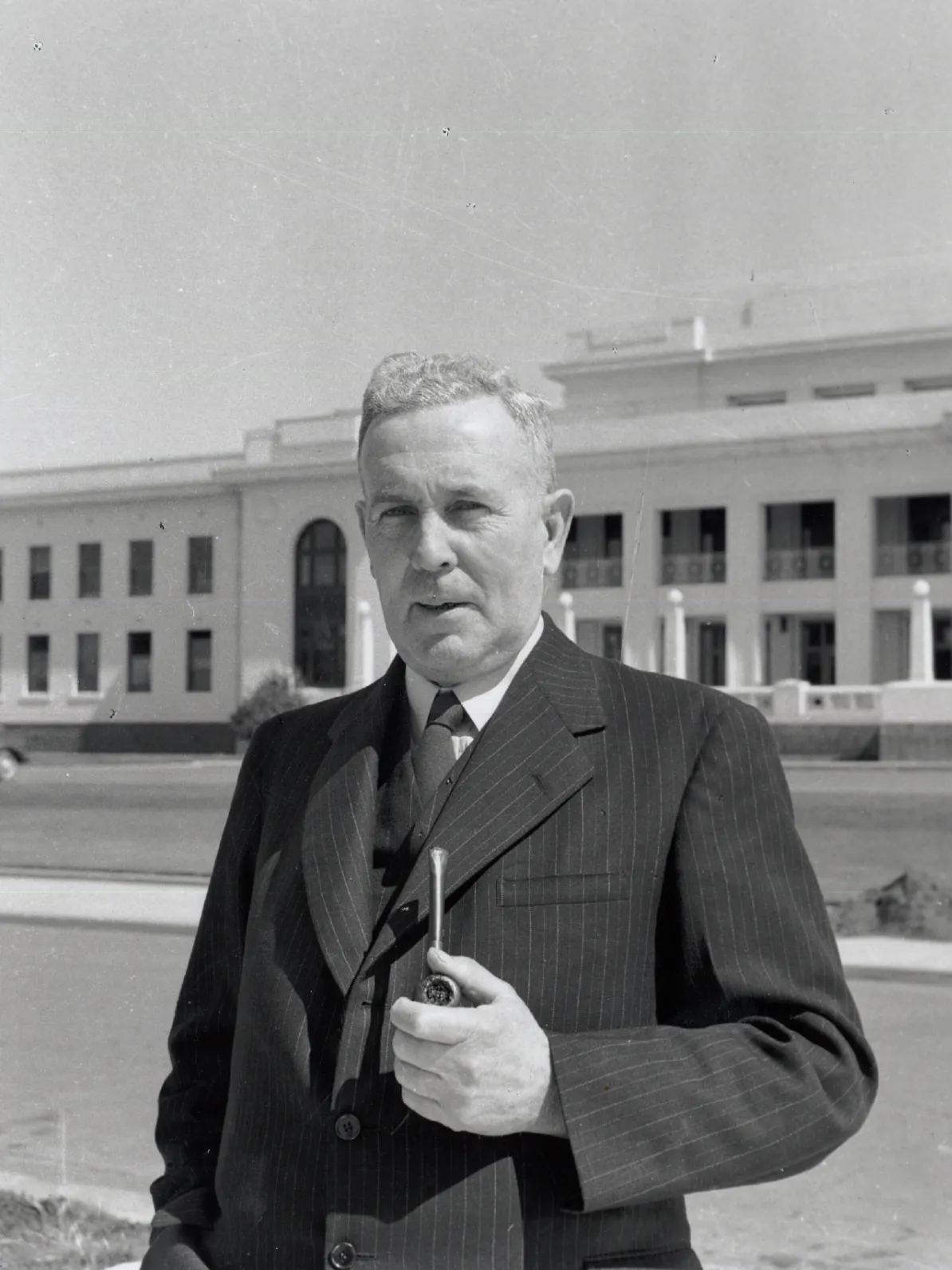 A black and white image of Ben Chifley standing in front of Old Parliament House in a suit with his pipe.