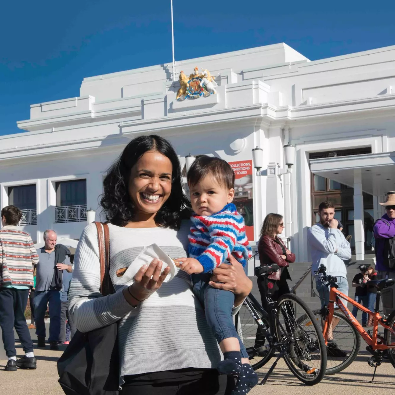 A mother holds her baby and smiles at the camera at the front of Old Parliament House. A row of people waiting to vote are behind her. She holds a sausage sandwich.