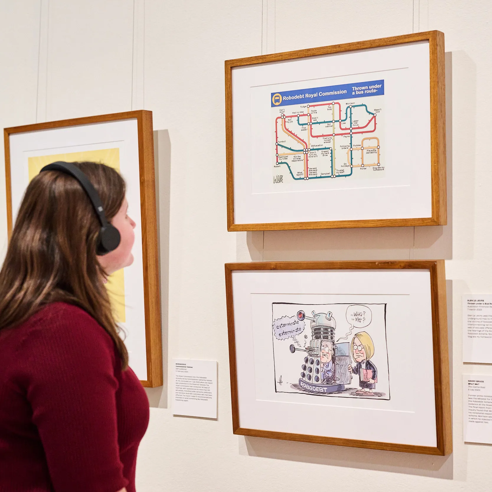 A woman wearing a dark red top and black headphones looks at framed cartoons with a caption featuring the Audio Description logo.