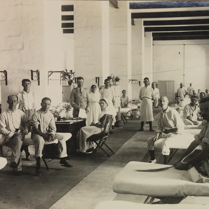 A surgical ward in Egypt during WWII with rows of beds with men looking at the camera. Earle Page is standing holding a piece of paper, next to a nurse. 