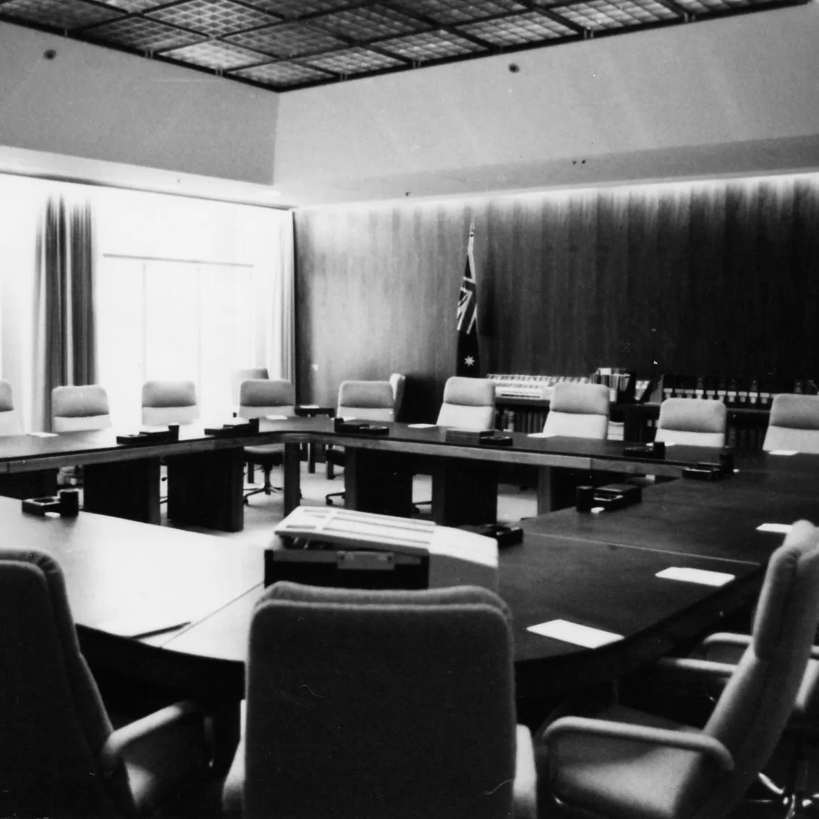 The Cabinet Room at Old Parliament House with a square table surrounded by chairs. Two Australian flags stand along a wood panelled wall.