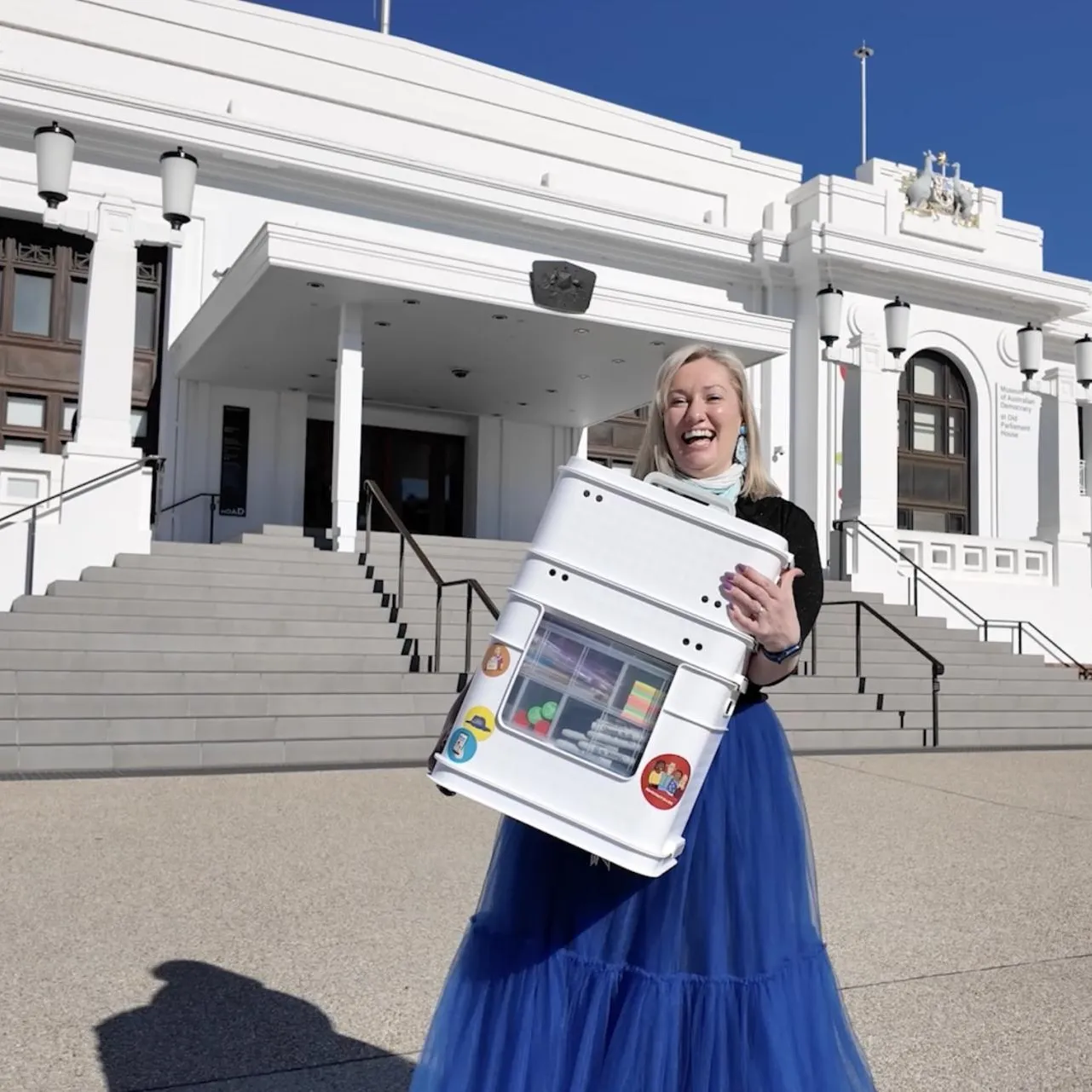 A woman in a blue skirt holds a box of activities outside Old Parliament House.