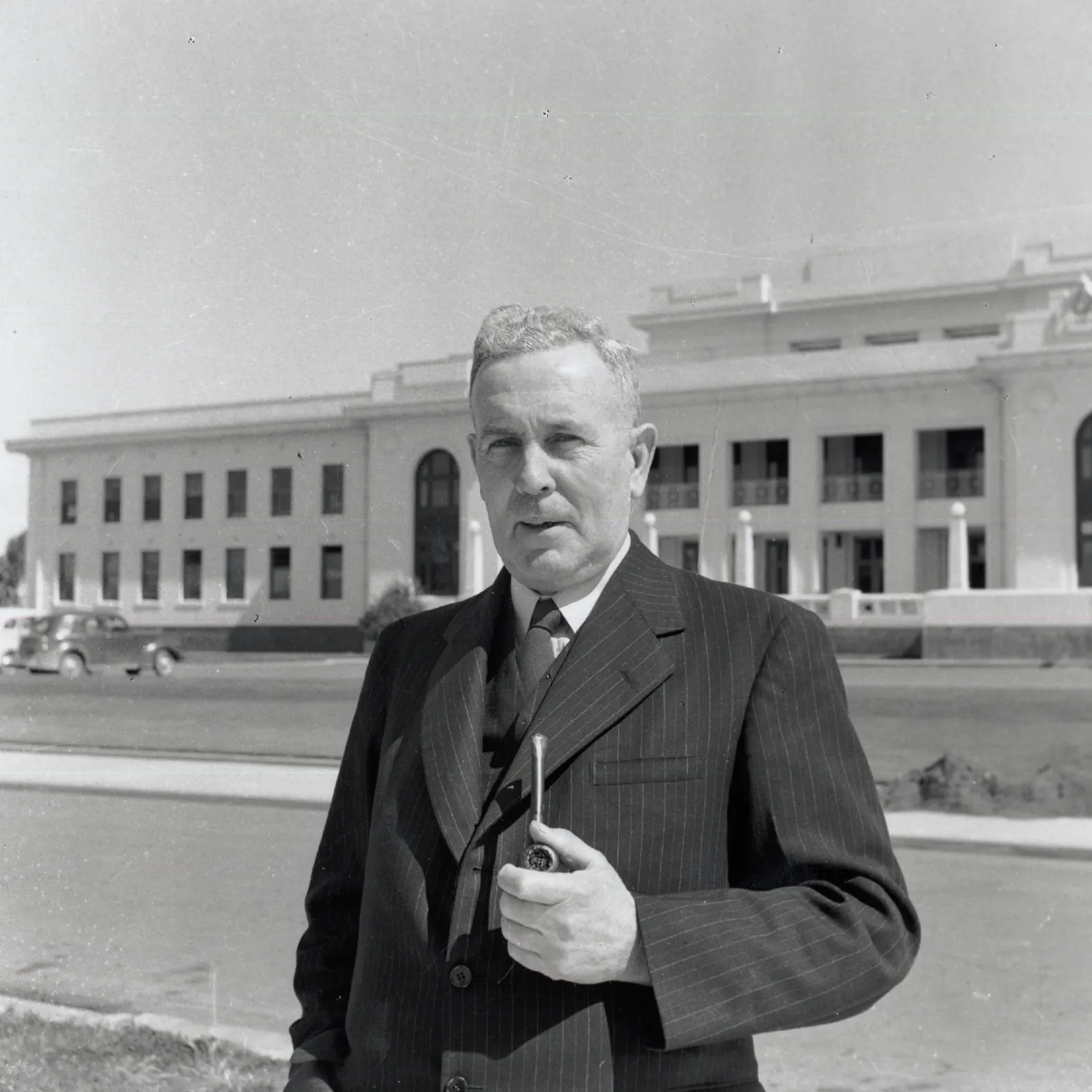 A black and white image of Ben Chifley standing in front of Old Parliament House in a suit with his pipe.