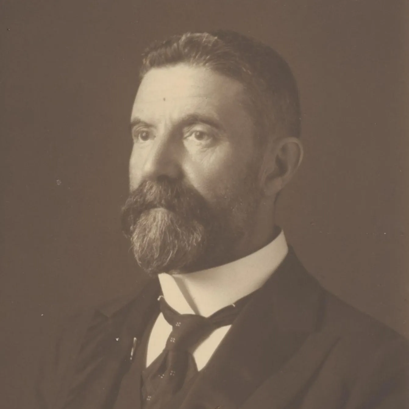A black and white headshot of Alfred Deakin. He's looking slightly left, in a suit and tie. He's got a short haircut with a well kept beard and moustache.
