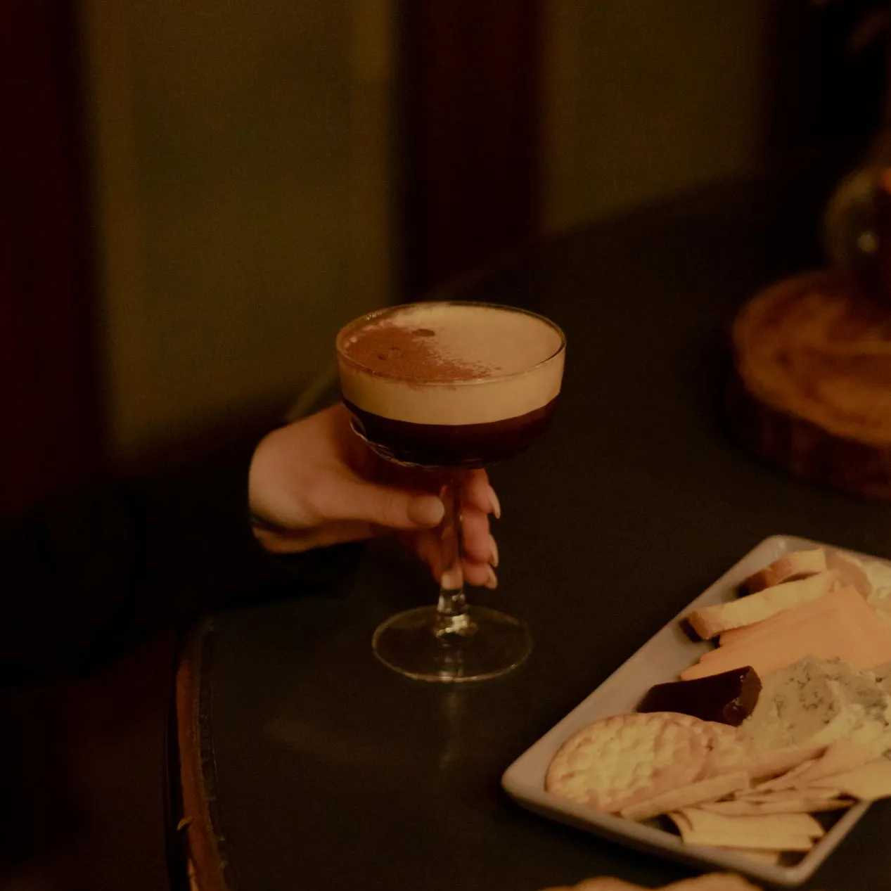 A hand holding an espresso martini on a table that also has a couple cheese plates.
