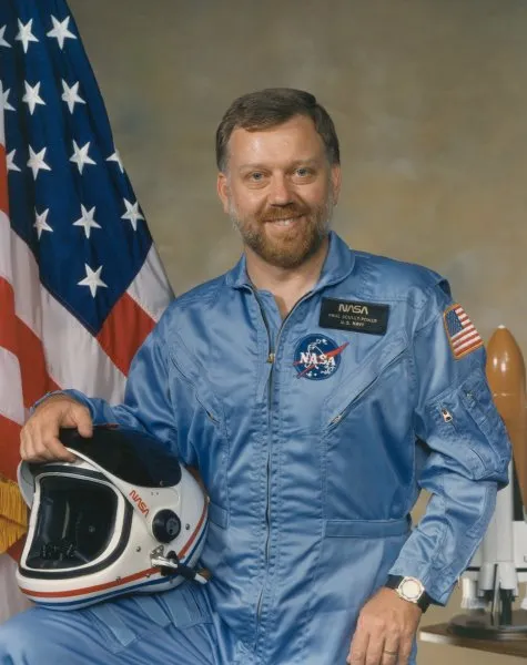 Astronaut Dr Paul Scully-Power wearing a blue NASA space suit with an American flag behind him.