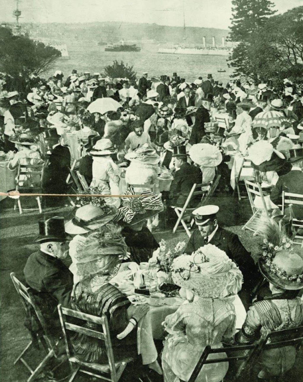 A large group of people enjoying tea at garden tables on the lawn, boats passing on the harbour in the background.