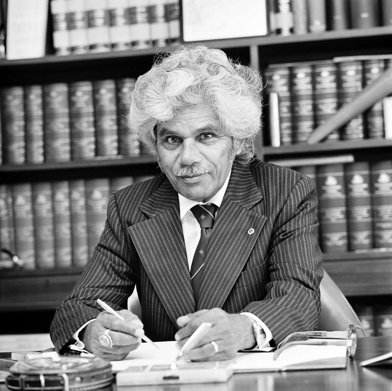 Neville Bonner sits at his desk, wearing a striped suit and tie, a pen in his hand, behind him a full bookshelf.