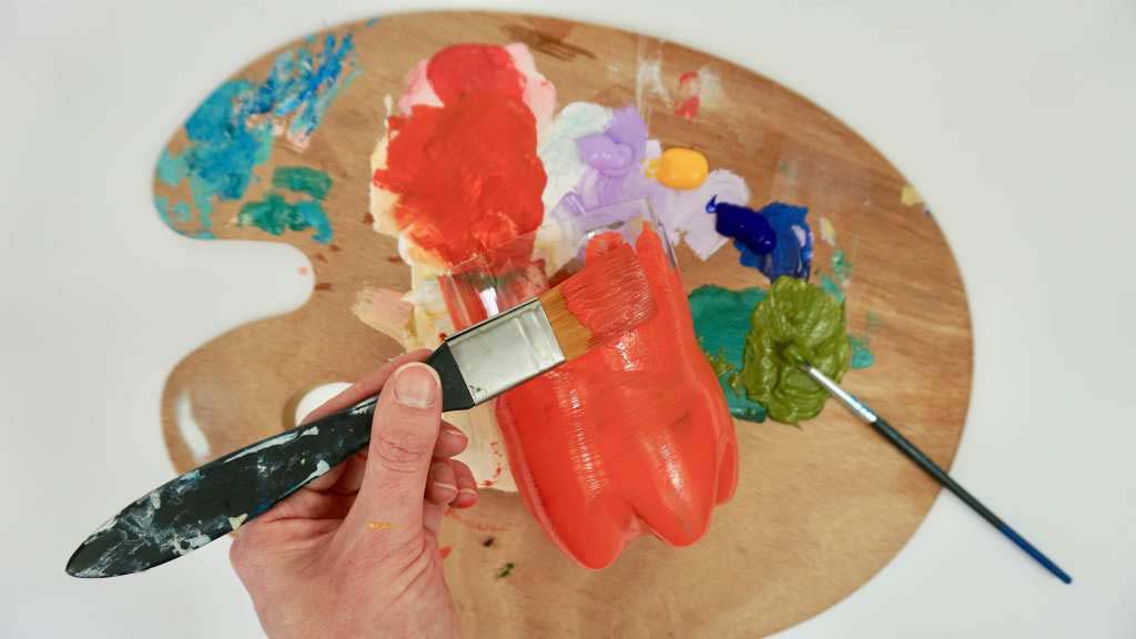 A close up of a hand holding a paint brush painting a plastic bottle red.