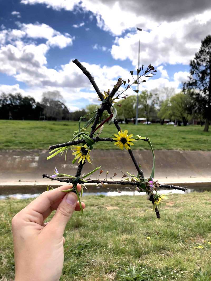 A close up of 4 sticks bound together to form a triangle shape with grass and flowers on it. 