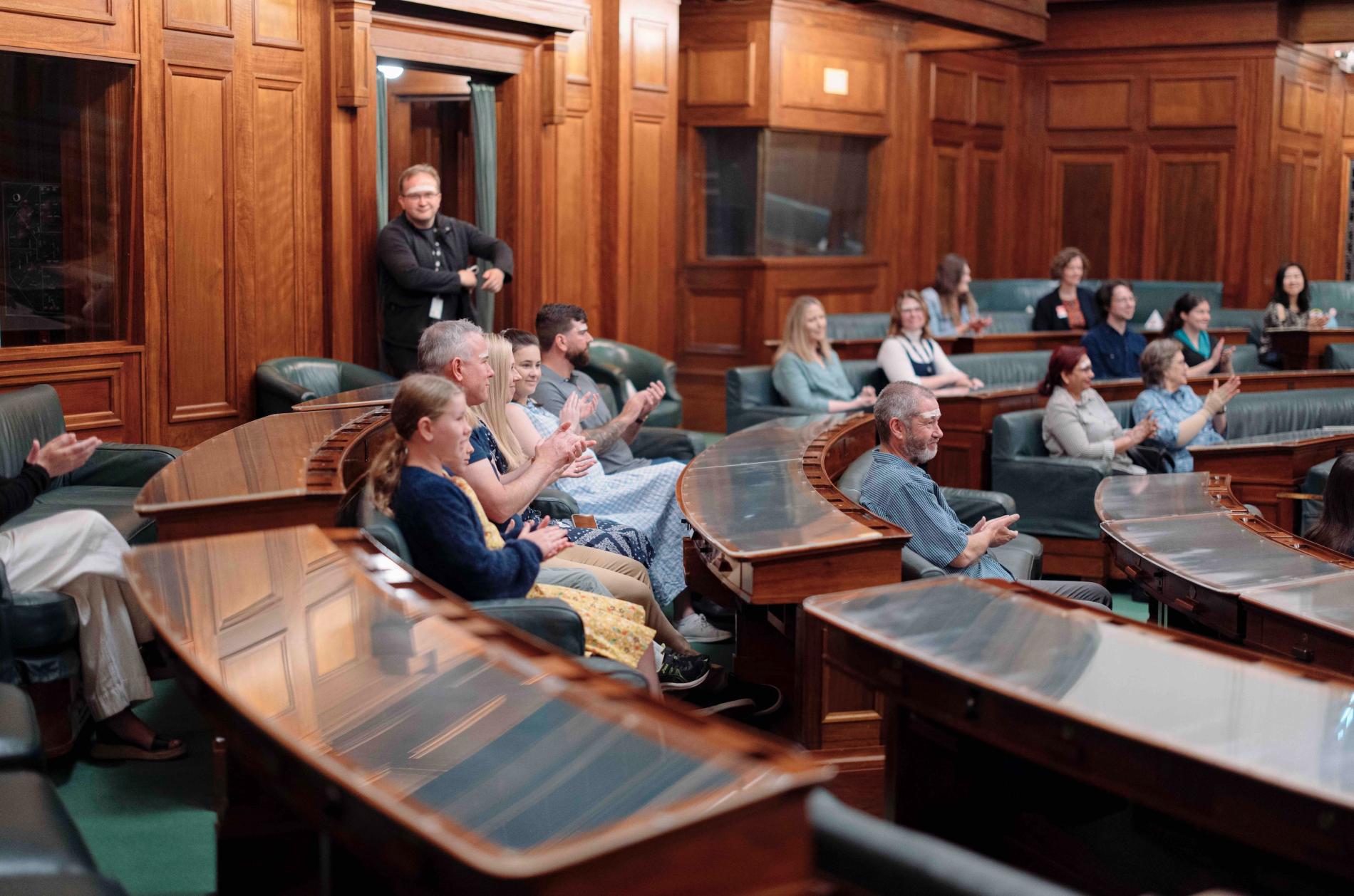 Visitors sit in green leather chairs with timber desks with glass coverings in a U-shape in the House of Representatives. 