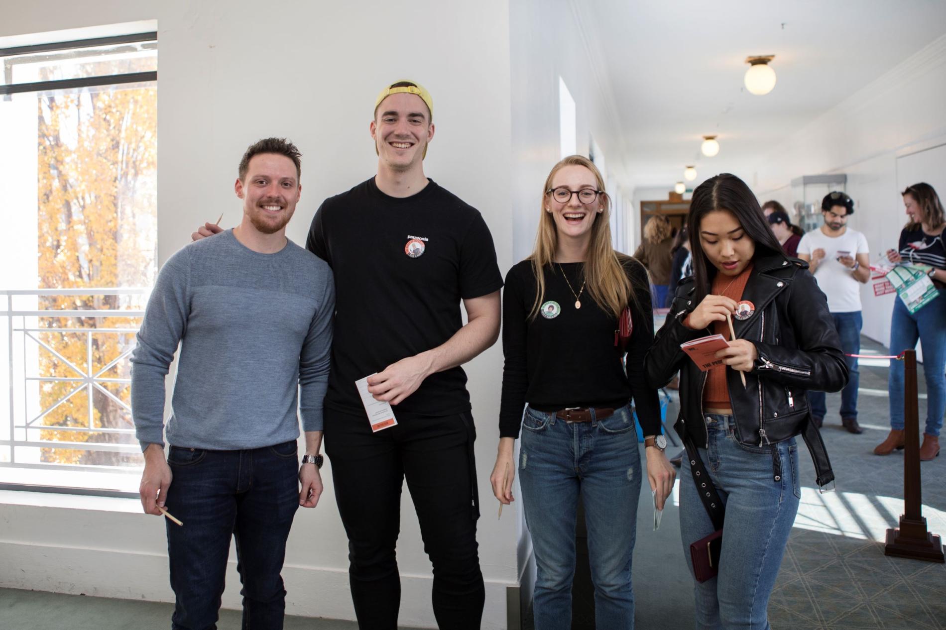 Two men and two women, stand in a row and a smile at the camera. They are wearing badges and holding voting cards. 