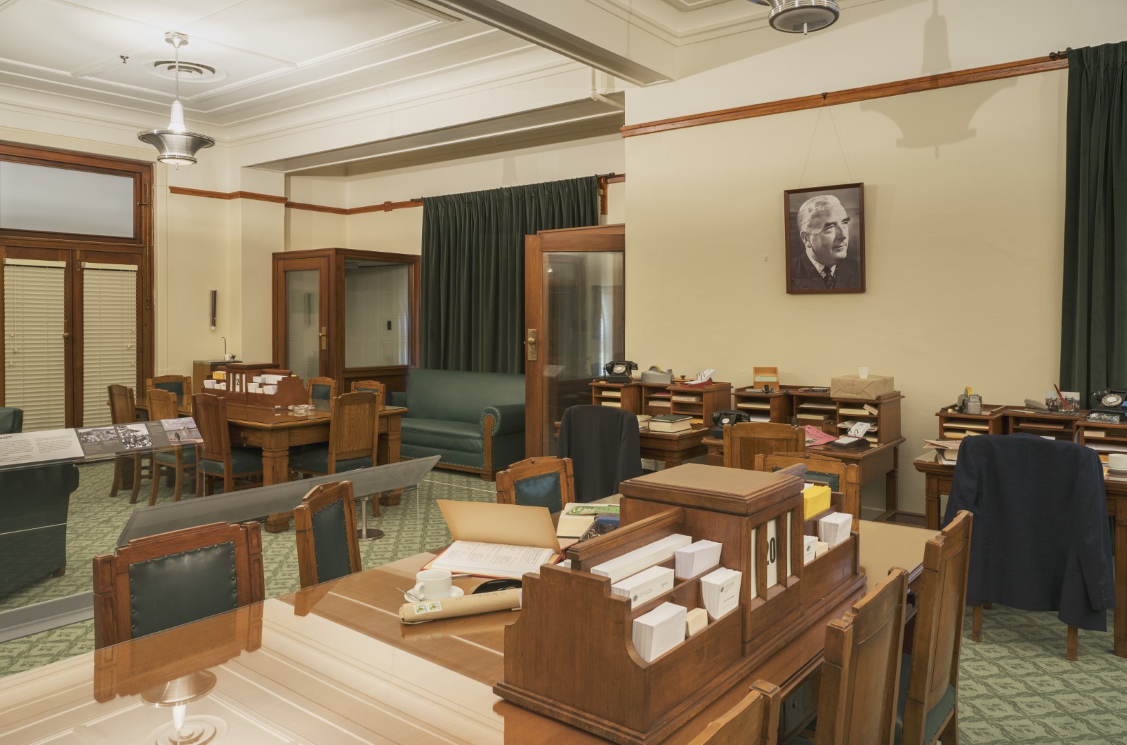 This colour photograph shows the Government Party Room re-created to evoke the atmosphere of 1964. There are two large meeting tables surrounded by chairs, three small desks, and two telephone booths. A large portrait of Prime Minister Robert Menzies is on one wall. The surfaces of the meeting tables and desks are covered with stationery, papers, telephones and ashtrays. 