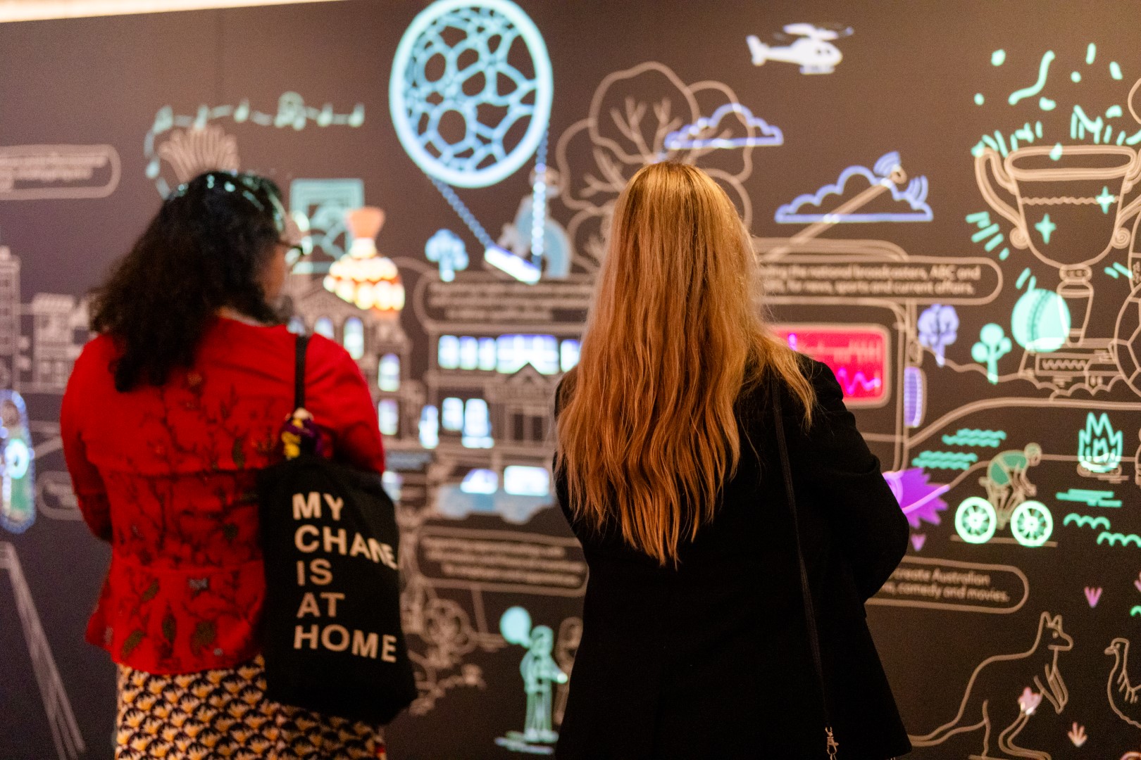 Two women look at a large wall installation of coloured lights and illustrations.