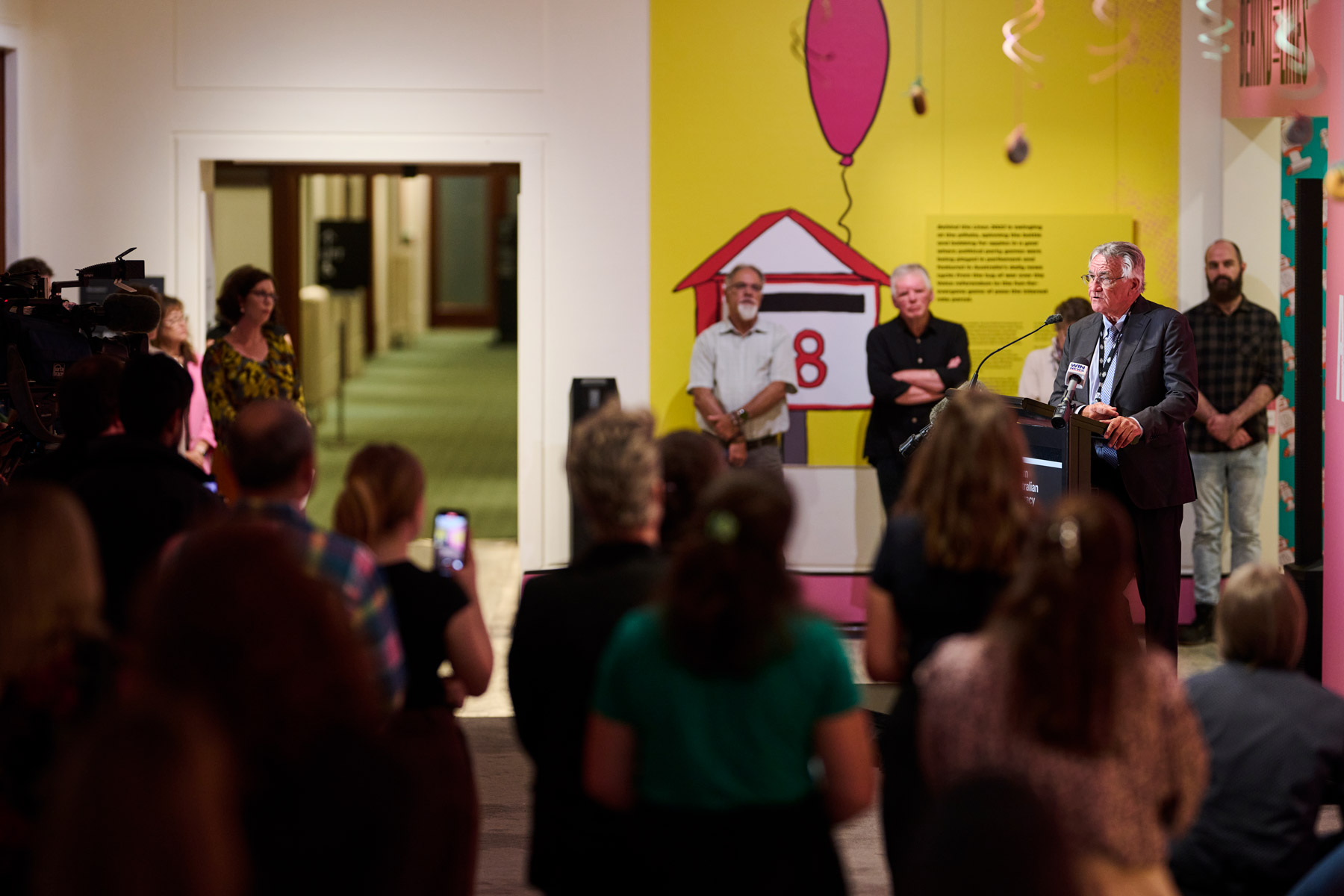 Barrie Cassidy stands behind a podium and speaks to an audience, some of whom are recording on photos and cameras. 