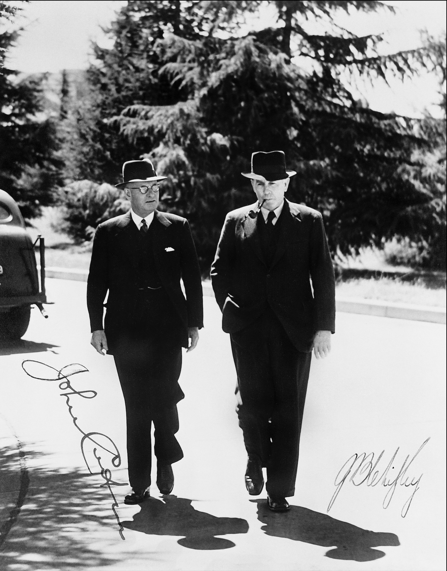 A black and white photo of two men in dark suits, wearing hats, one with glasses and the other with a pipe in his mouth walking. There are two signatures on the photo. One reads John Curtin, the other reads Ben Chifley. 
