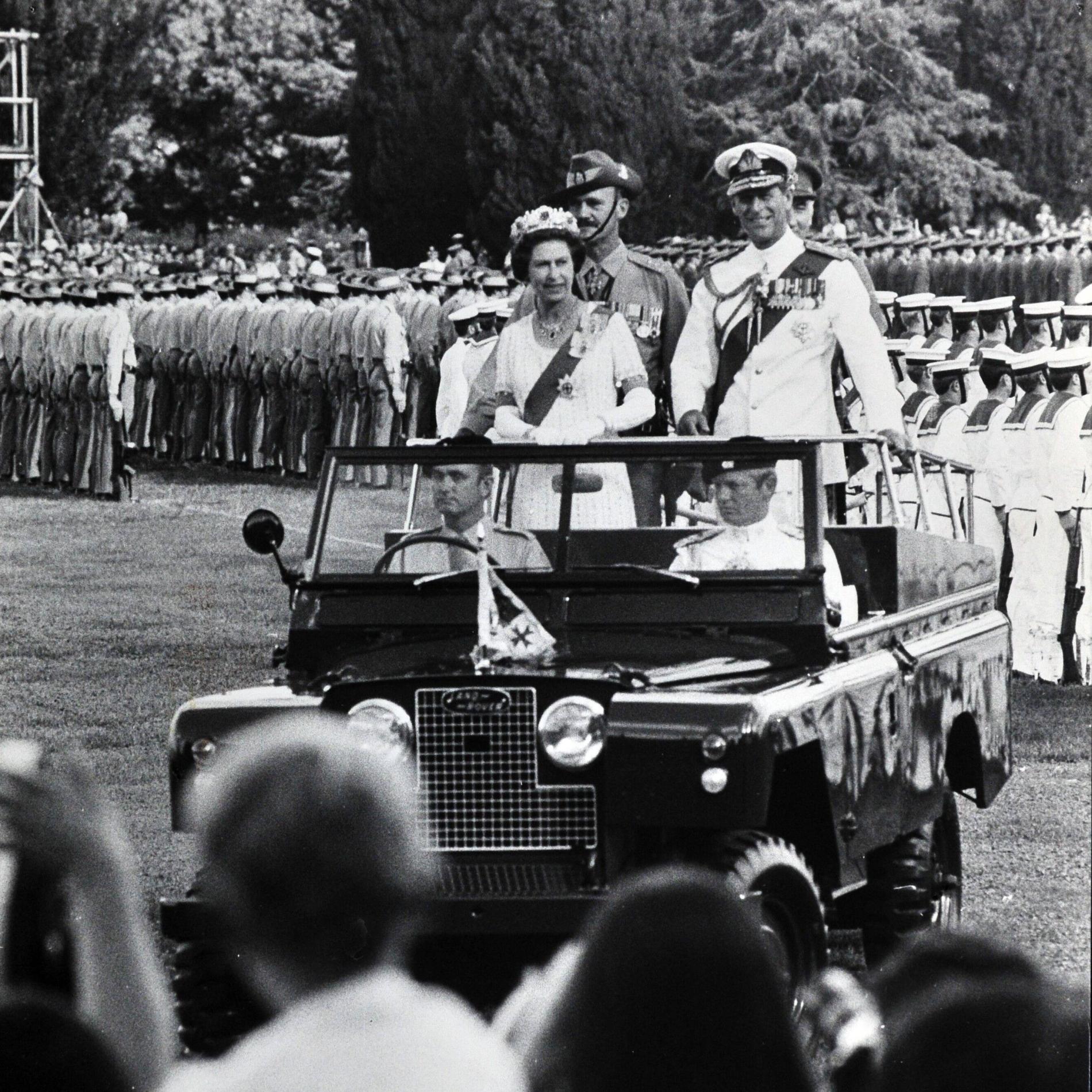 Queen Elizabeth II and Prince Philip stand on the back of an open-topped car, passing troops standing in formation.