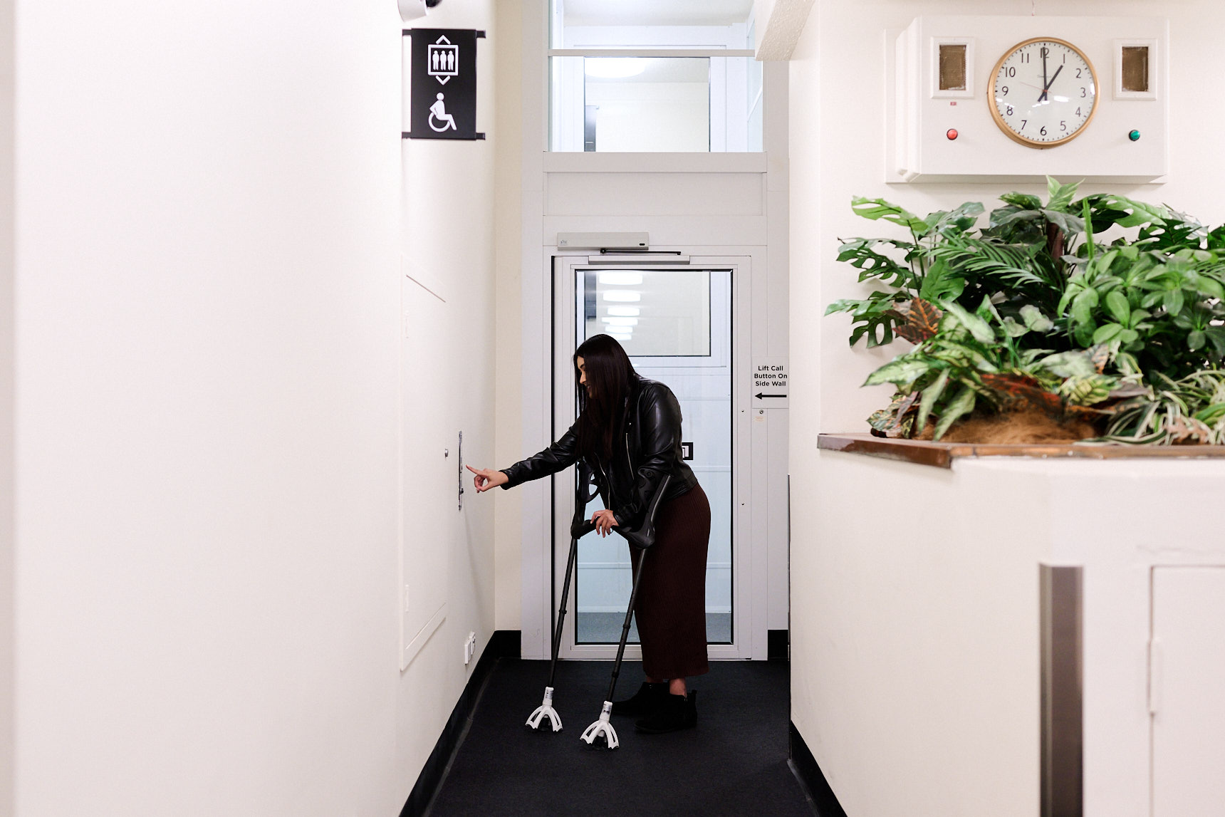 A woman on crutches presses the lift call button for the accessible lift in a corridor of MoAD.