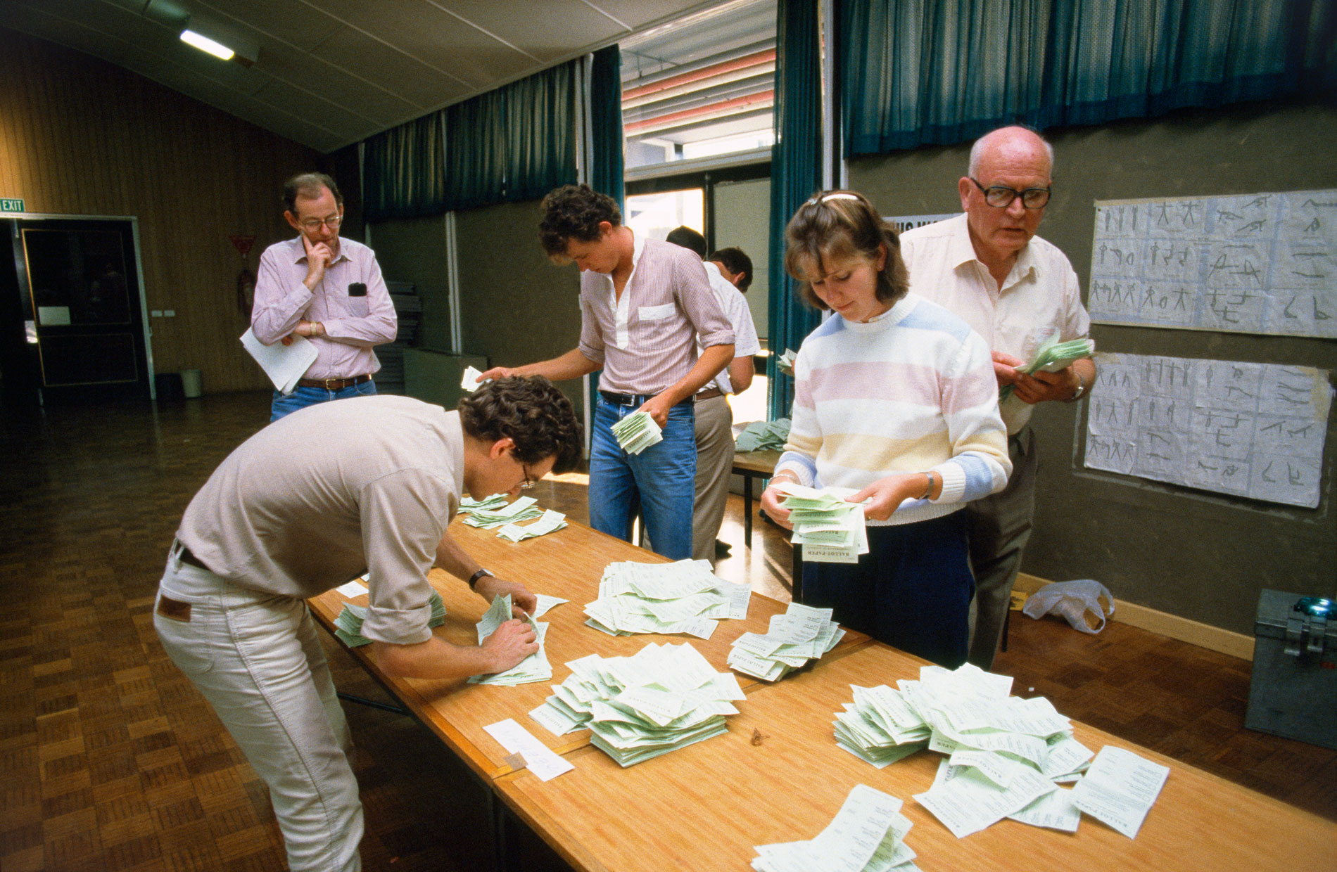 Men and women count and sort green ballot papers on a wooden table in the 1984 election.