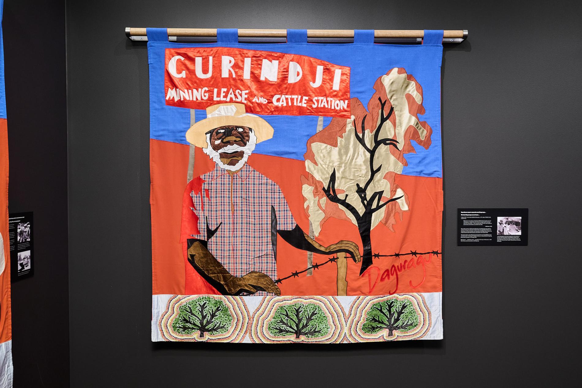 A Gurindji freedom banner depicting a First Nations person. The banner hangs in the exhibition on a black wall in Old Parliament House.