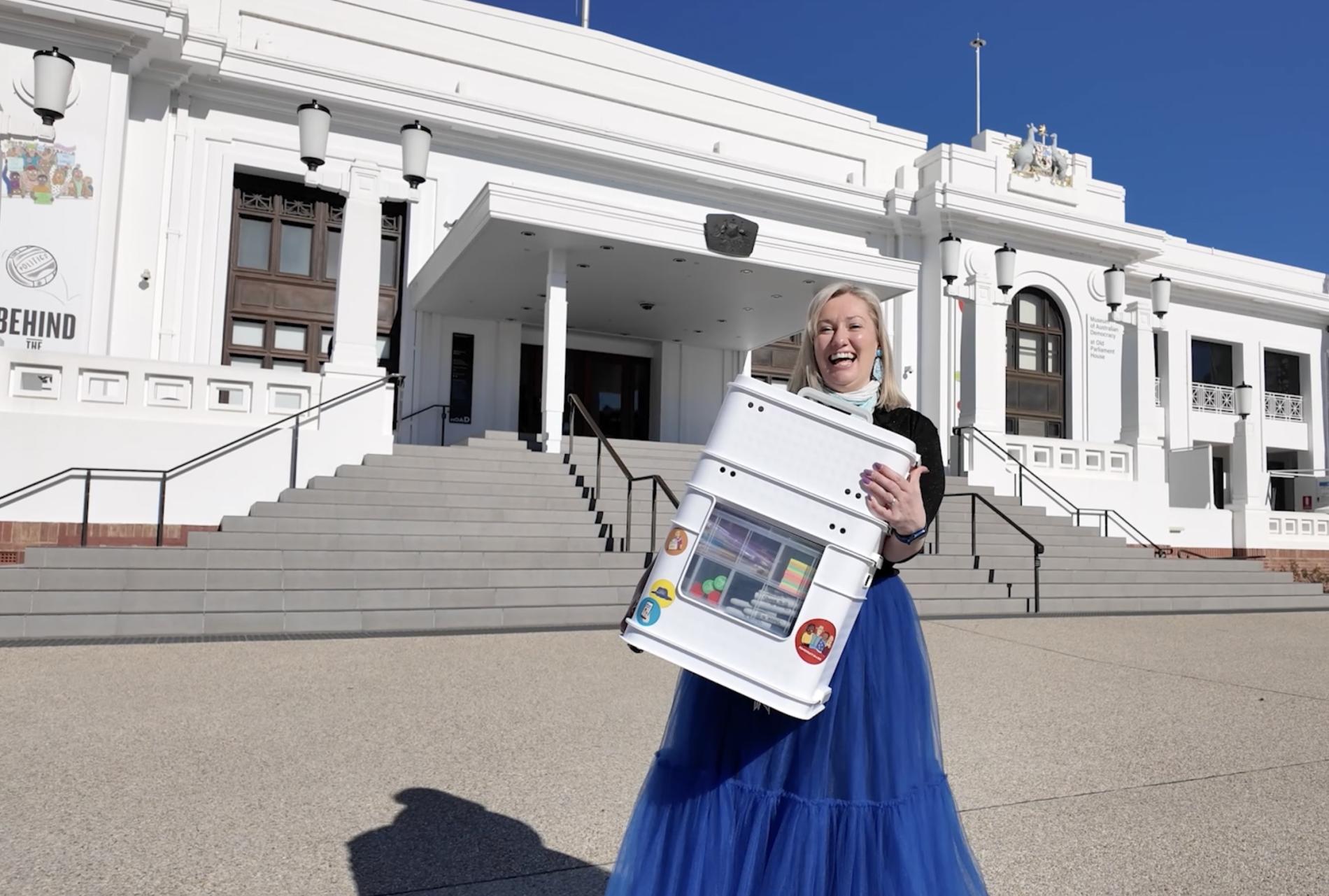 A woman in a blue skirt holds a box of activities outside Old Parliament House.