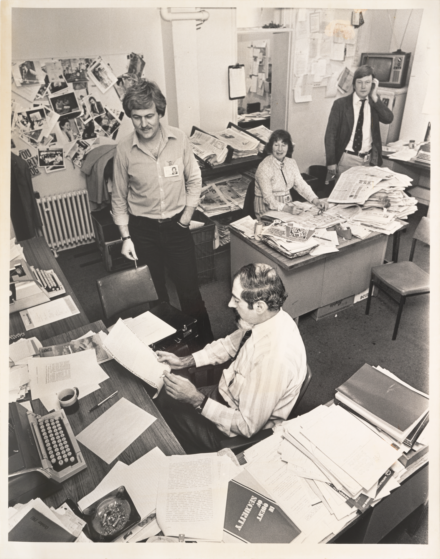 Press Gallery journalists in a small room covered in newspapers, photographs and notes. In the front, there's a man sitting at a typewriter smoking a cigarette, reading over a transcript.