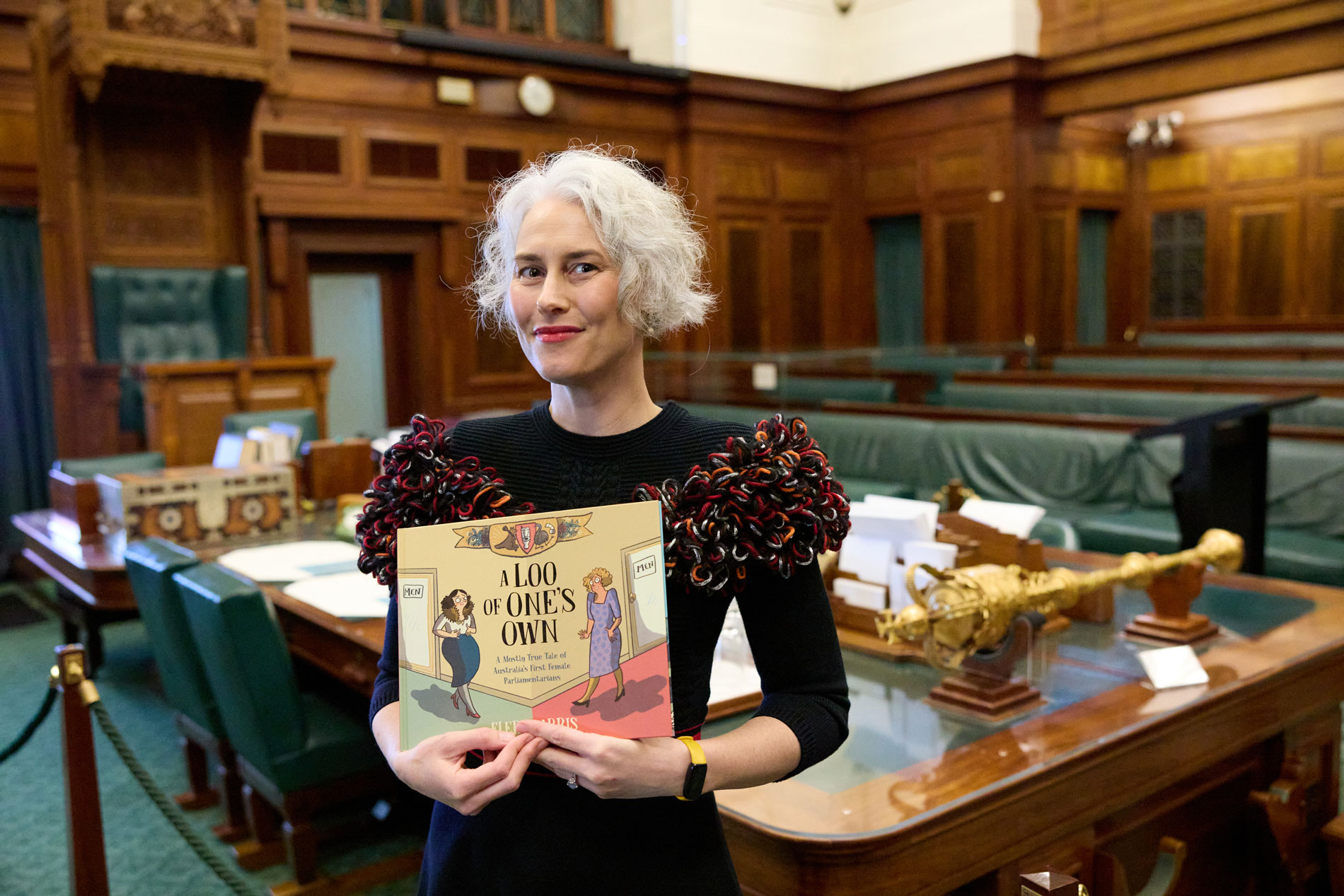 Author Eleri Harris holds a book standing in the green Senate Chamber of Old Parliament House.