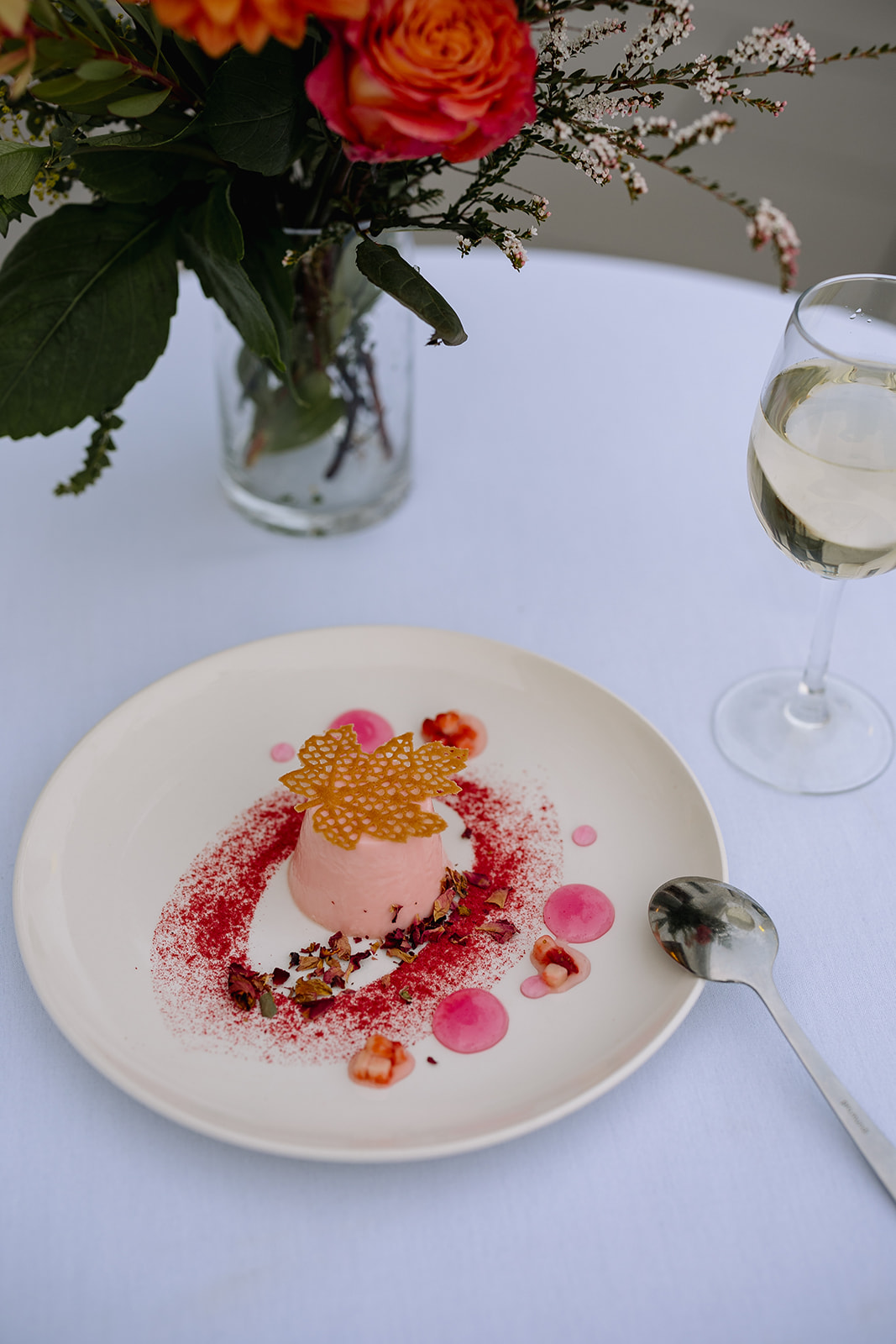 A pink dessert laid out delicately on a plate. In the corner, there's a wine glass with some white wine inside.