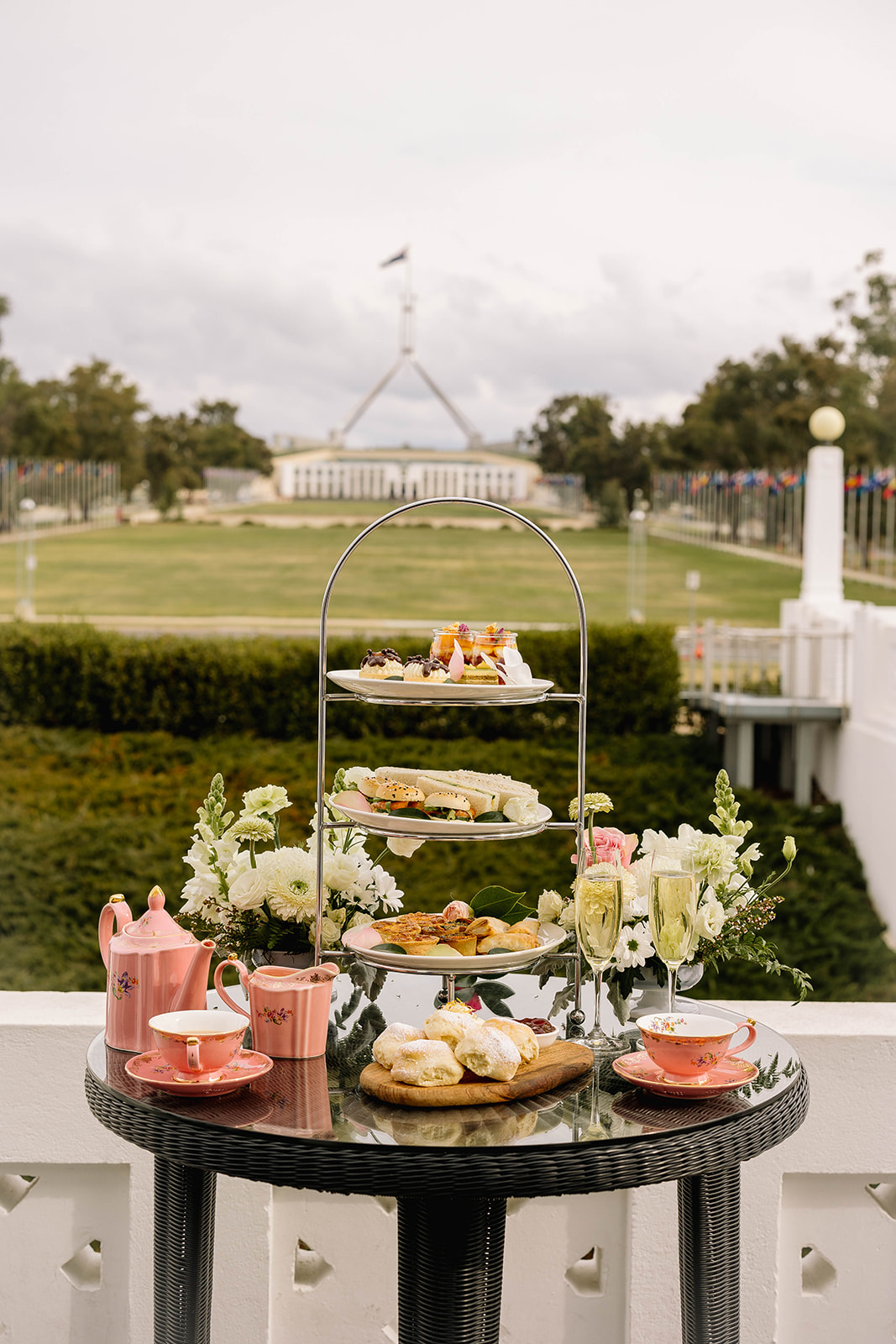 A tiered cake stand, each layer contains different food like sandwiches, pastries and bread rolls. In the background, you can see Australian Parliament House.