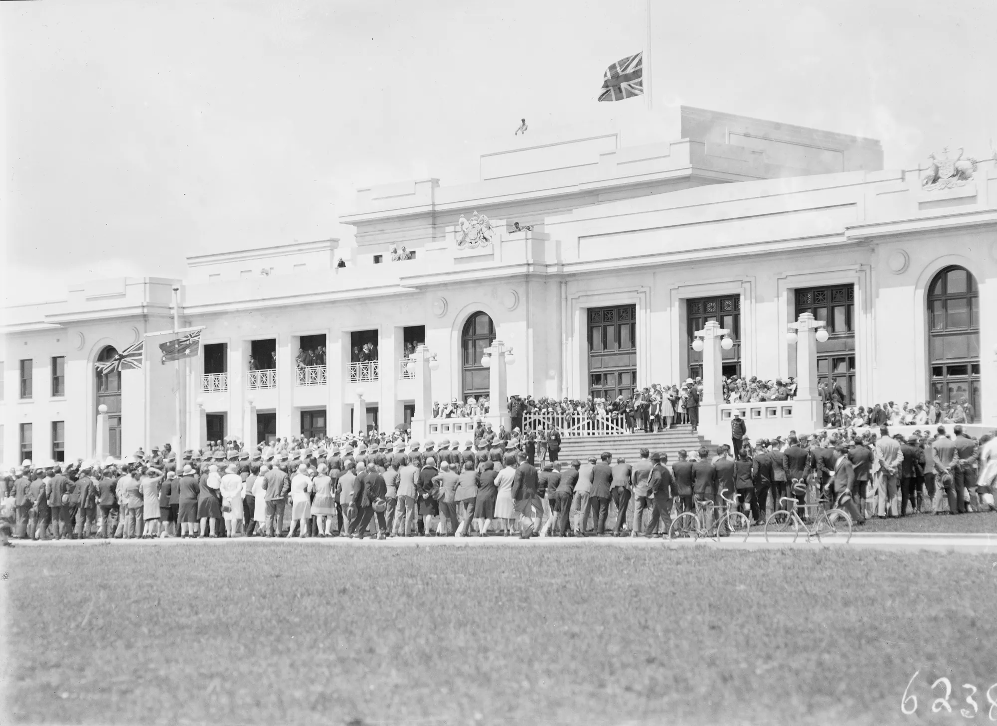 Black and white image of the front of Old Parliament House with a crowd of people of gathered at the foot of the stairs. The building is white with columns and an Australian flag flies at the top. 