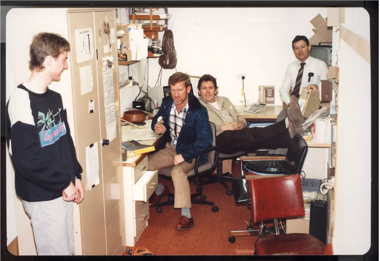 This amateur colour photograph gives us a peek into the small, crowded and messy office occupied by the Telecom employees at Old Parliament House. The office is crowded with desks, chairs, bookshelves and a cupboard. A young, smiling Scott Cadden stands to the left in jeans and sweatshirt, Neil Baker sits at a desk holding a telephone handset and dressed in trousers, checked shirt and casual blue jacket. He has red hair and a beard and was known as ‘Bluey’. Seated behind Neil is a smiling Jeff Wilke with hi