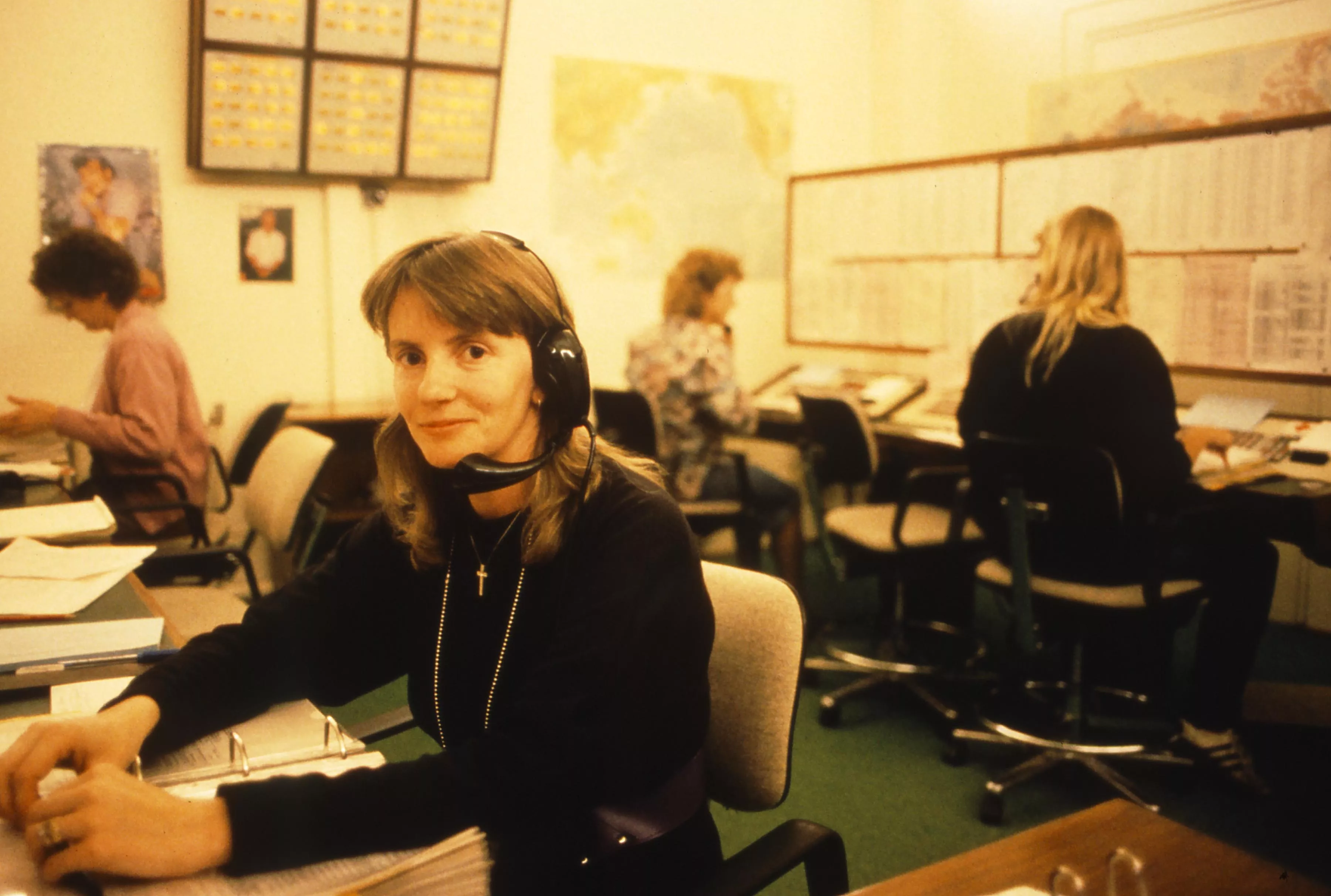 This professional colour photograph lets us view the switchboard operators’ office in Old Parliament House in 1988. In the foreground, a young woman with a gentle smile looks directly into the camera. She has shoulder length brown hair and a fringe and is wearing a black long-sleeve top. She is wearing a headset with headphones and a mouthpiece and her hands are resting on a thick binder. There are three other women seated at desks checking listings and handling telephone calls. The room has a large pinboar