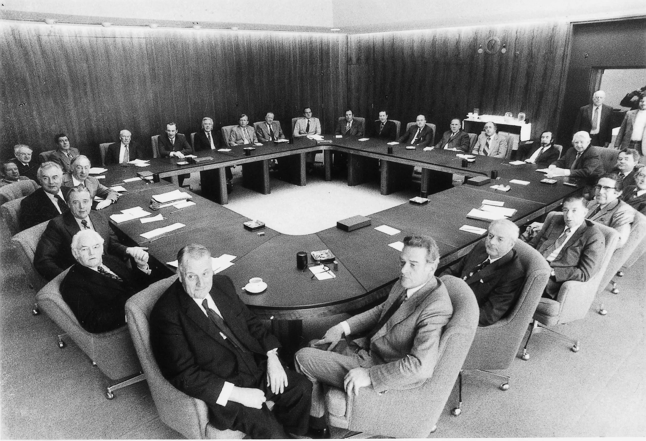 This black and white photograph is taken from one corner of the timber panelled Cabinet Room making the square table appear as a diamond. The all-male Cabinet are looking at the camera and are all in standard business attire.  On the far right three men stand in the doorway to the room. The table is relatively neat with papers in stacks and smoking accessories in neat groupings as the meeting has not yet begun.  