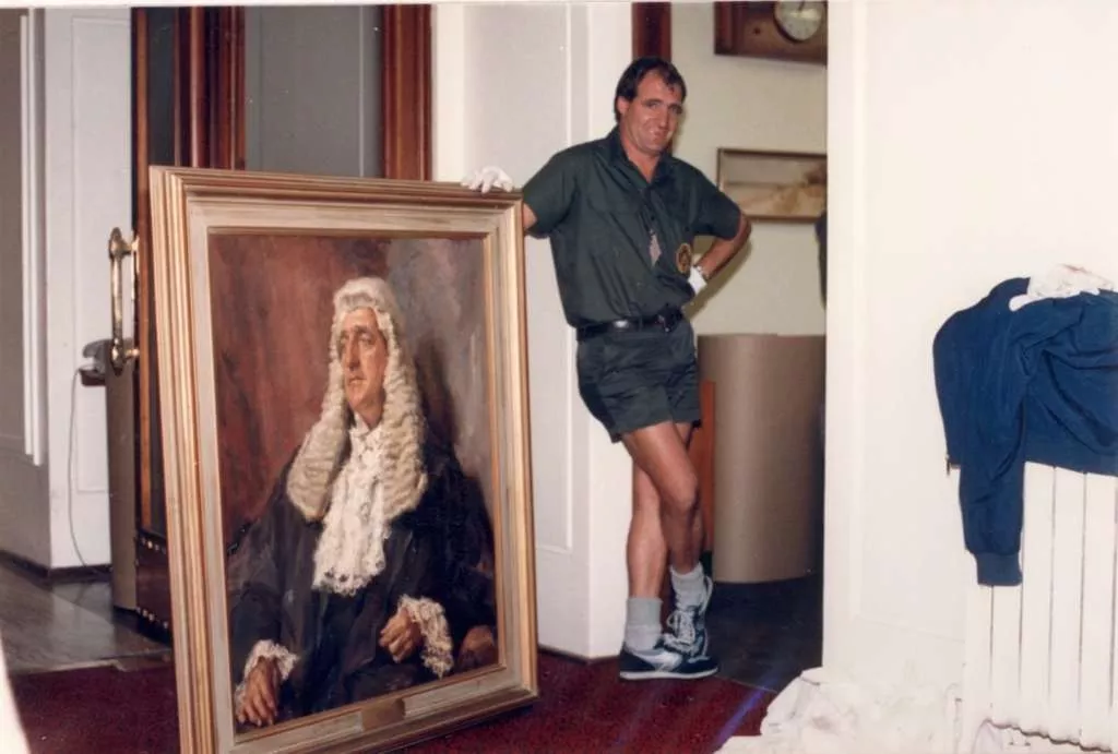 A removalist wearing white gloves posses next to a portrait painting of a main wearing the President of the Senate's wig, black gown and lacy collar.