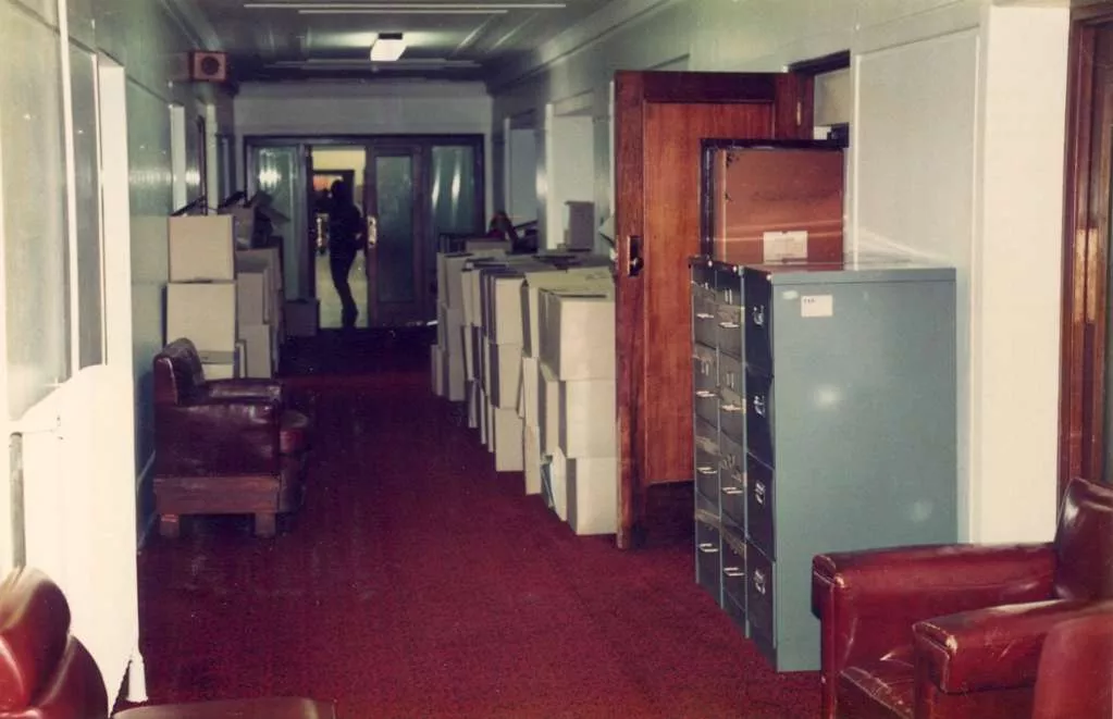 A red-carpeted corridor, lined with large cardboard boxes, filing cabinets and red leather furniture.