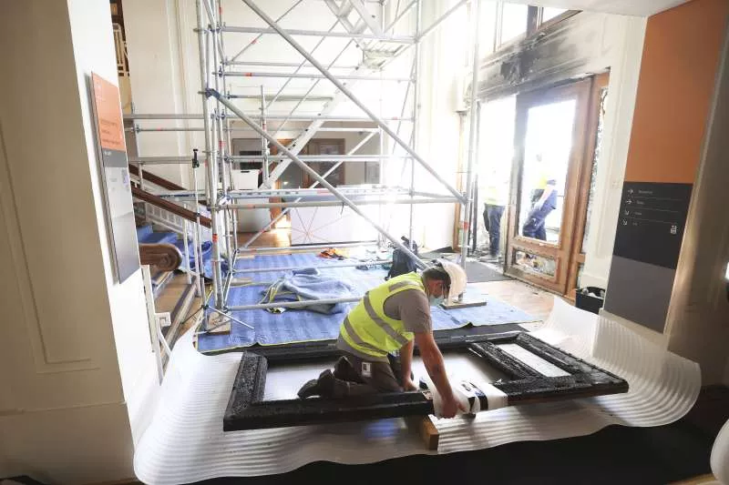 Scaffolding is set up inside the smoke damaged entrance of Old Parliament House, and a man kneels on the ground wrapping a burnt door frame.