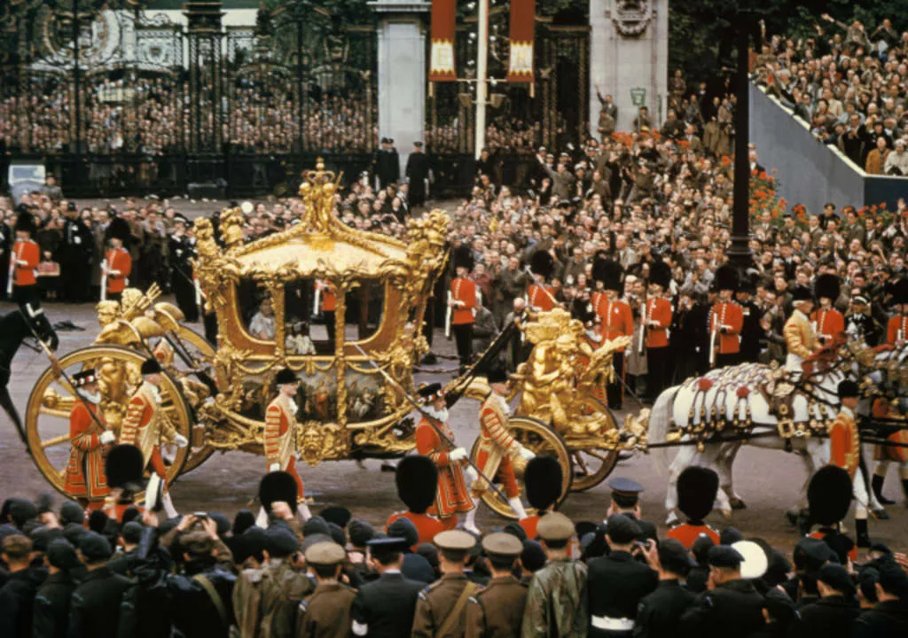 Queen Elizabeth II sits in an ornate golden carriage, pulled by horses down a crowded street.