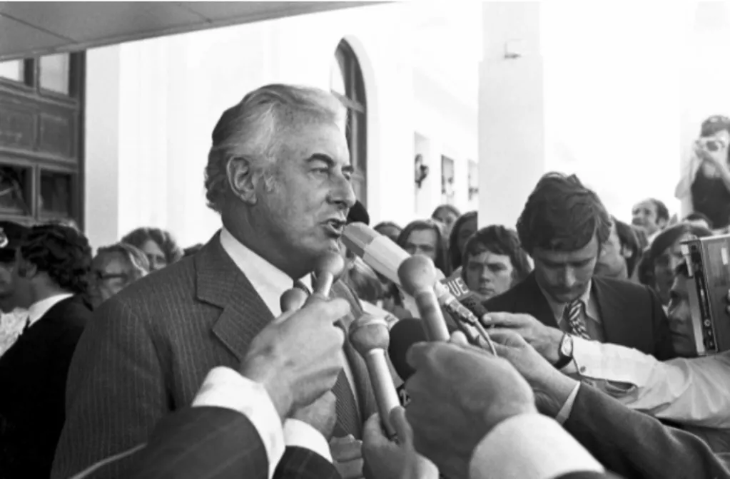 Black and white photograph of Gough Whitlam speaking to a large group of people holding microphones to his face, in front of parliament house immediately following the Dismissal.