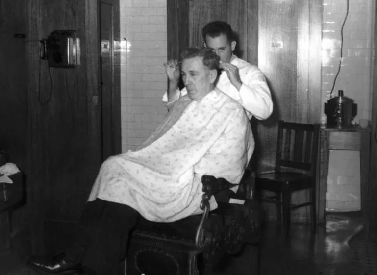 Ben Chifley sits in a chair with a large white patterned cloth covering his clothes as Cecil Bainbrigge cuts his hair with scissors.