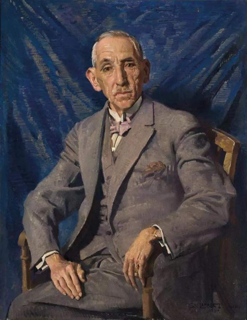 Former prime minister Billy Hughes in a grey suit, with a pink bowtie, sitting in a wood chair in front of a dark blue curtain.