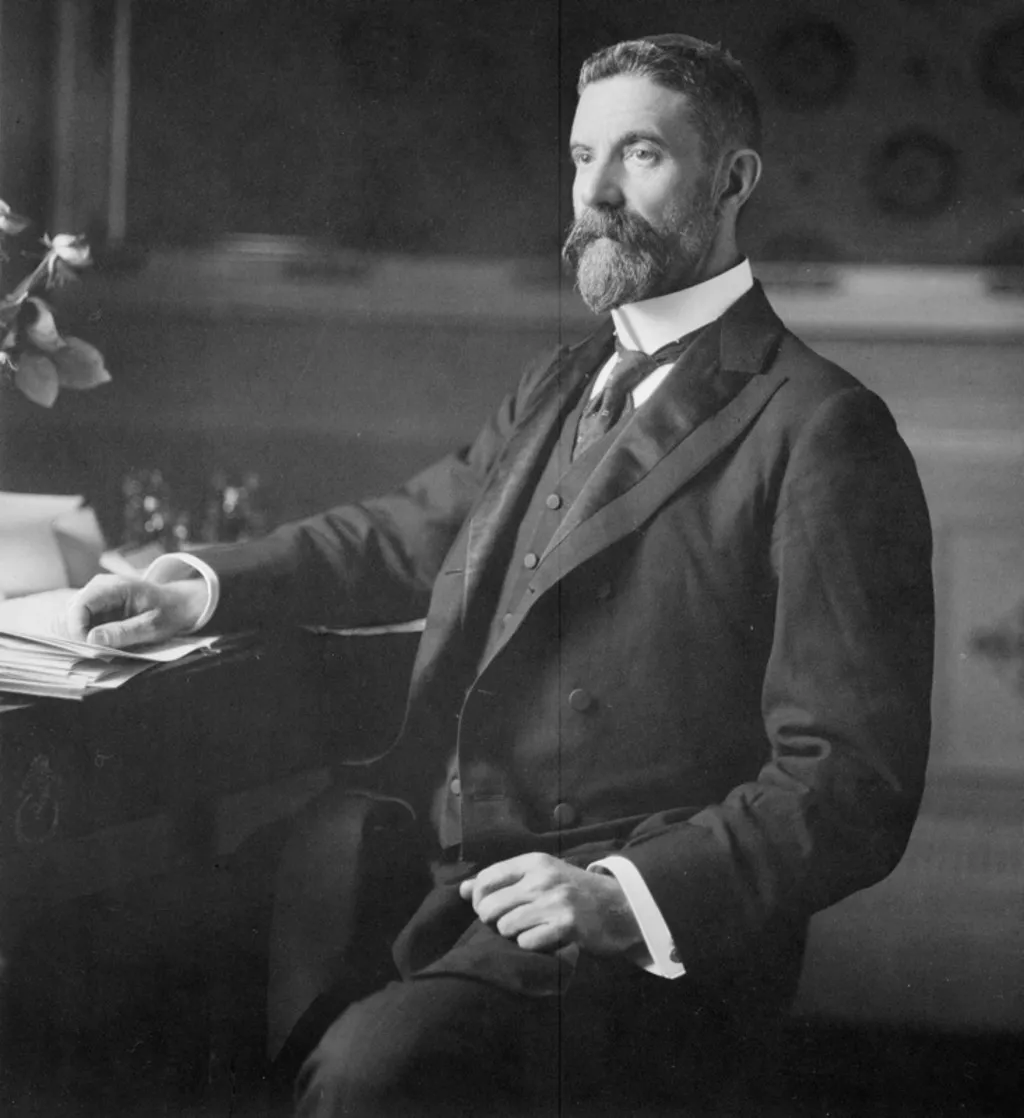 Alfred Deakin, wearing a three-piece suit and seated at a desk covered in paper documents.