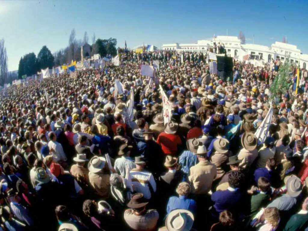 A very large crowd of farmers, many wearing hats and waving banners, rallying in front of Old Parliament House.