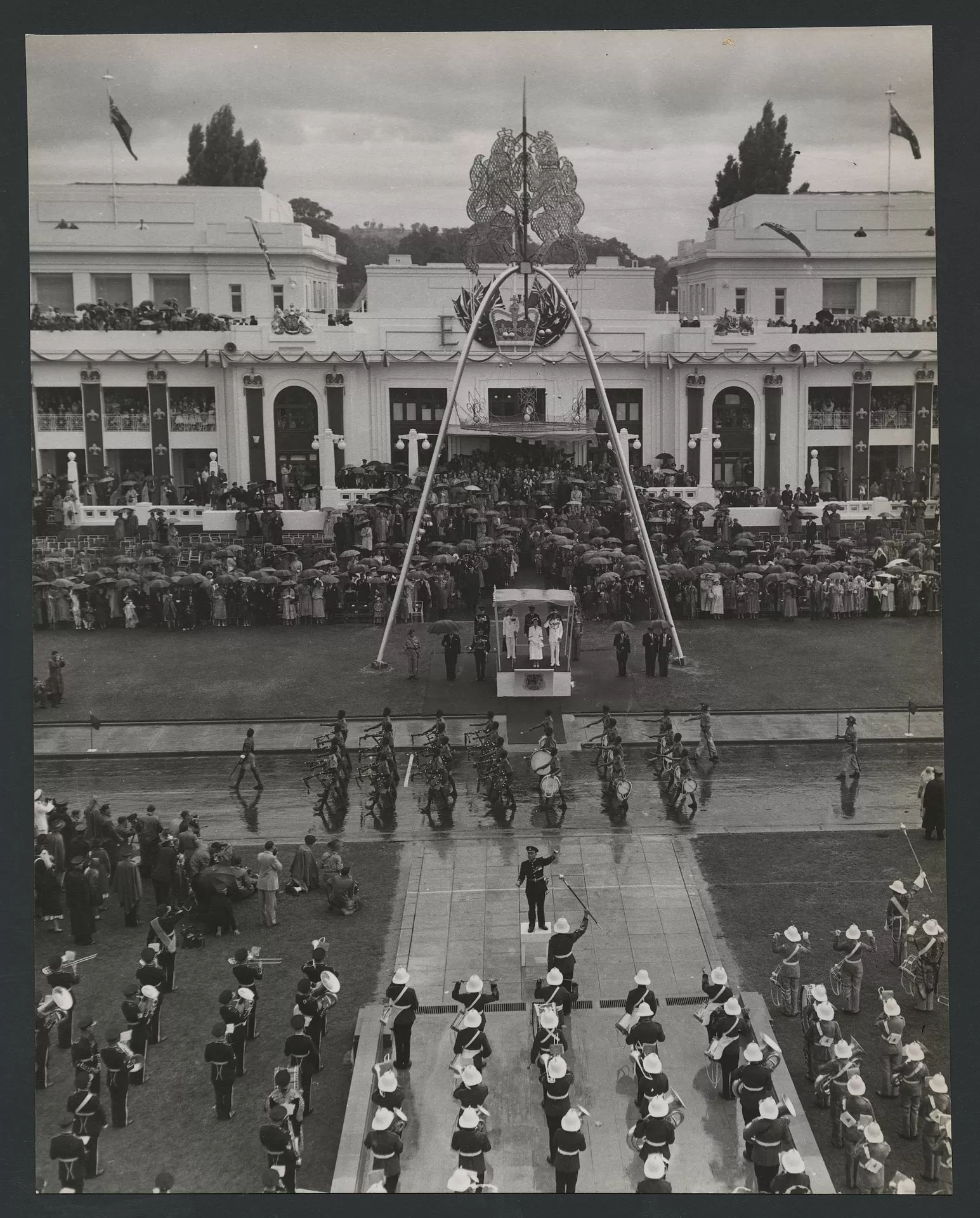 Overhead view of Queen Elizabeth standing in a podium out the front of Old Parliament House, surrounded by a crowd holding umbrellas as troops march past.